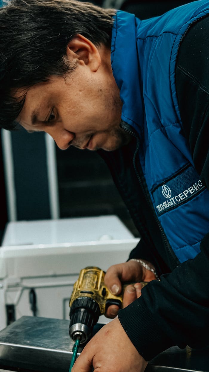 Technician in blue jacket using a power drill indoors for maintenance.